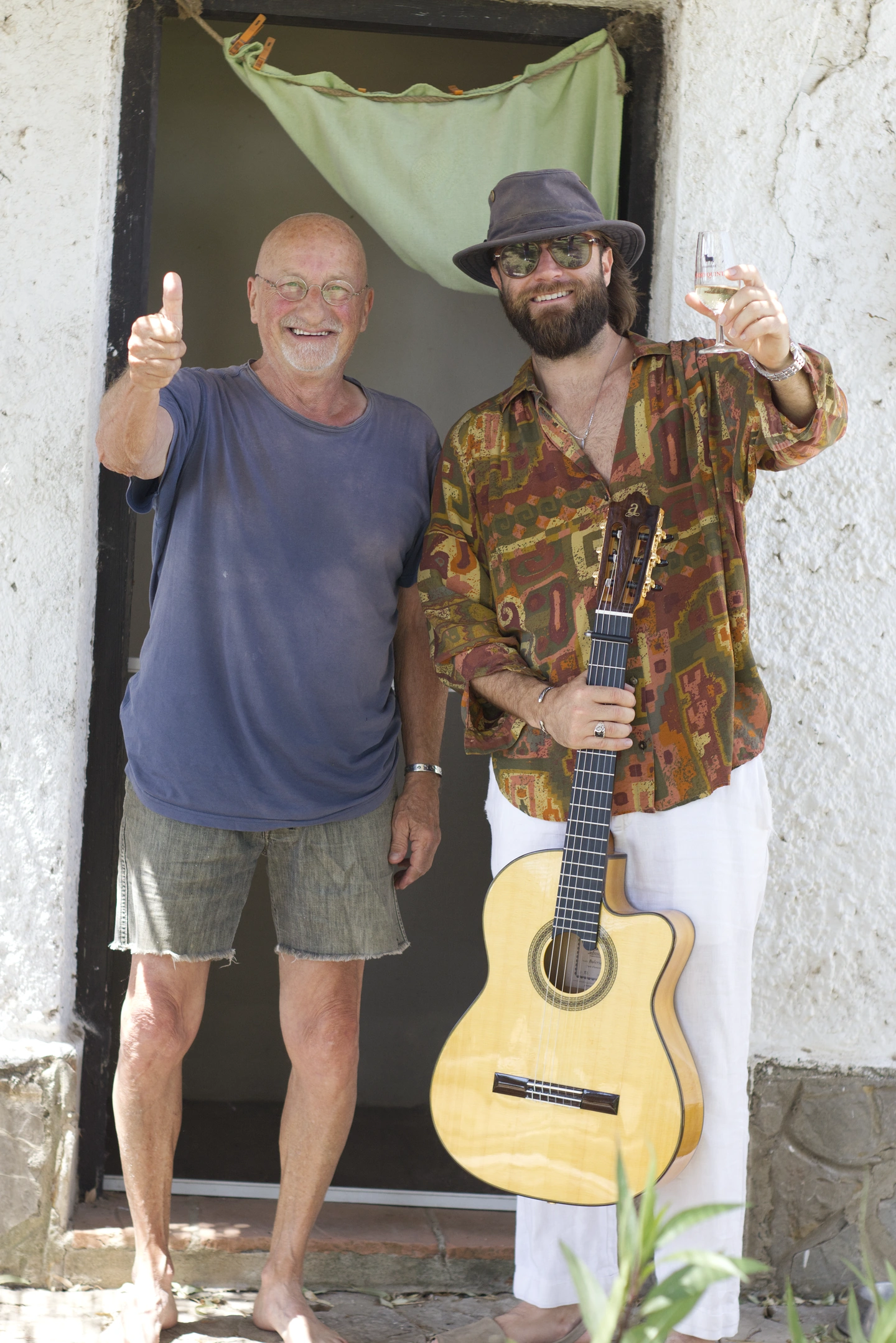 El Higüanero sostiene su guitarra y sonríe junto a otro hombre en la puerta de una casa blanca.